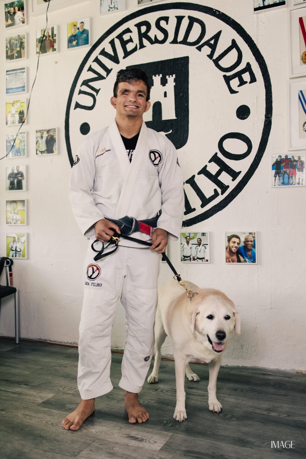 Carlos Alvarez, smiling and barefoot in a white jiu-jitsu gi with a black belt, standing with his cream Labrador guide dog in front of a Universidade Gama Filho emblem painted on an academy wall lined with framed photos