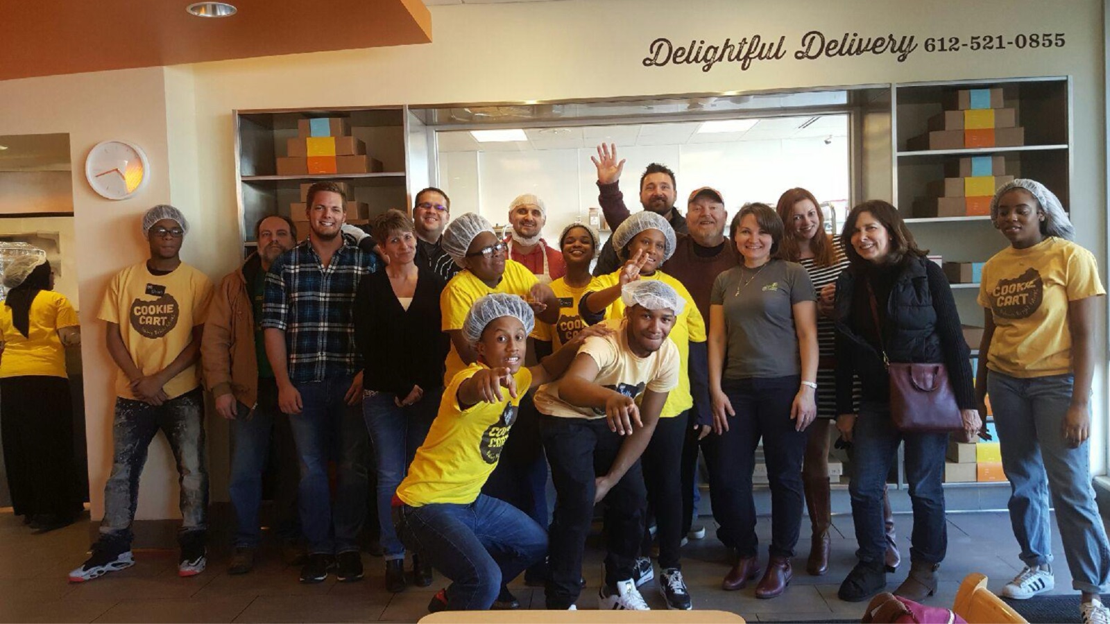Homestead Road team members and Cookie Cart teen bakers in yellow t-shirts posing together inside the Cookie Cart bakery in Minneapolis