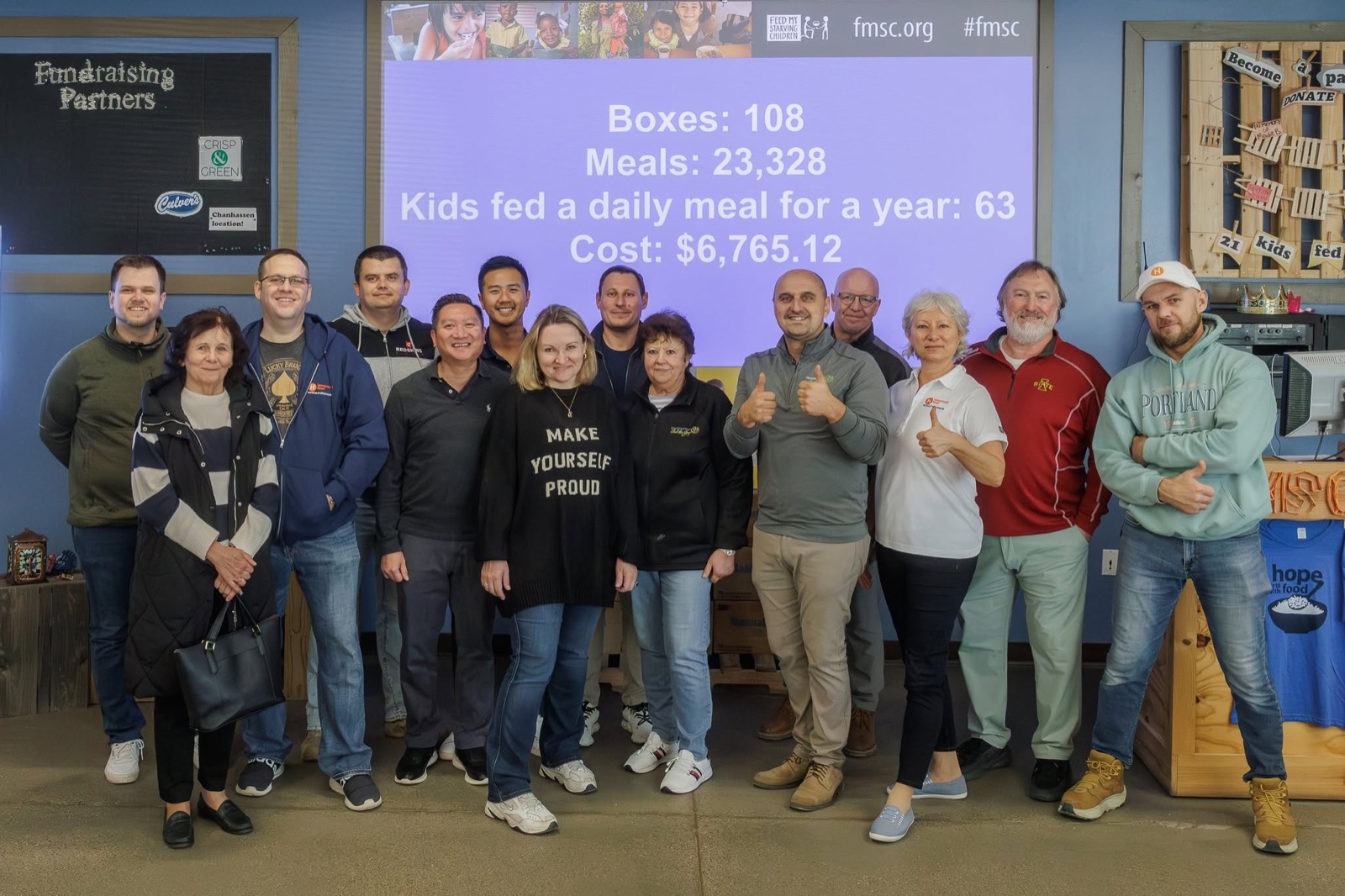Homestead Road volunteers giving thumbs-up at Feed My Starving Children in Chanhassen, Minnesota, in front of a screen showing 23,328 meals packed during their session