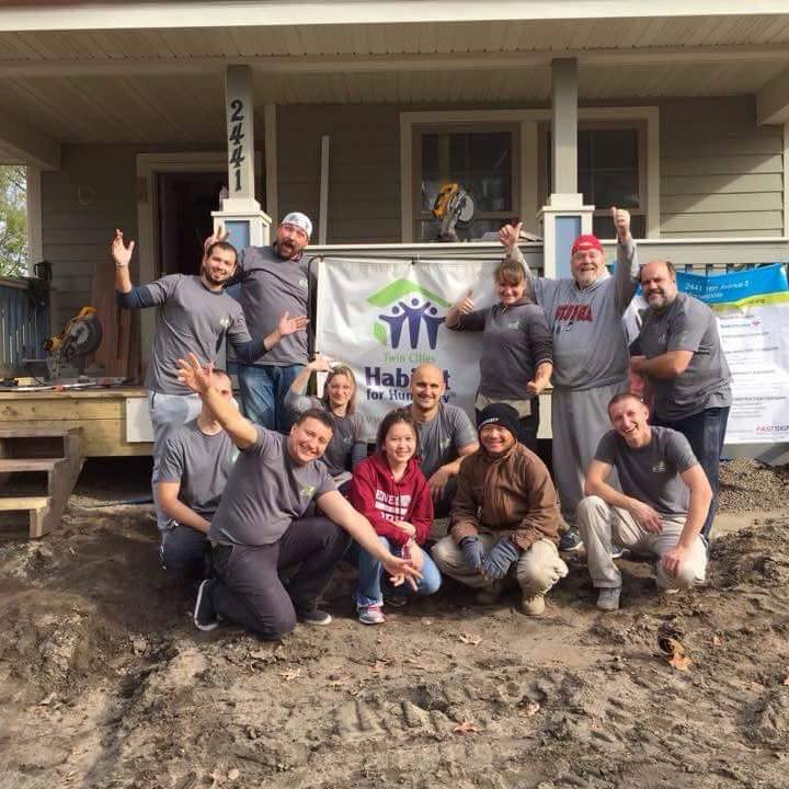 Homestead Road volunteers celebrating with raised arms in front of a Twin Cities Habitat for Humanity banner at a home under construction on a volunteer build day